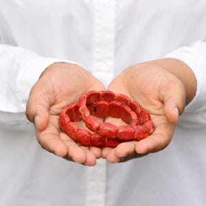 Puede incluir: Dos pulseras de coral rojo se sostienen en manos abiertas contra una camisa blanca. Las pulseras están hechas de cuentas rectangulares ensartadas. Las pulseras son de un color rojo vibrante y las cuentas tienen una forma ligeramente irregular.