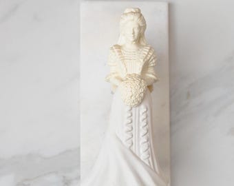 May include: A cream-colored statue of a woman in a long dress, holding a bouquet, mounted on a rectangular base. The statue has intricate details in the clothing and hair, and is set against a marble background.