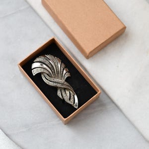 Op de afbeelding: Zilverkleurige Art Deco broche met een waaiervormig ontwerp, versierd met kleine, sprankelende steentjes. De broche wordt gepresenteerd in een bruine kartonnen doos met een zwart interieur, het deksel staat iets open. Een accessoire in vintage stijl.
