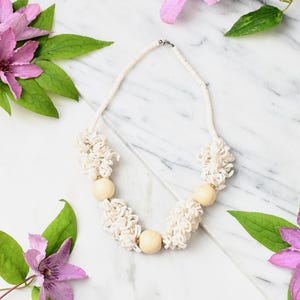 Op de afbeelding: Een ketting met een streng van kleine witte schijfkralen, afgewisseld met drie gladde, ronde, lichtbeige houten kralen en vier clusters van getextureerd, opgerold wit materiaal. De ketting is voorzien van een zilverkleurige metalen sluiting.