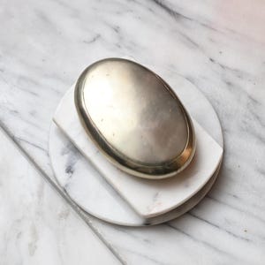 Puede incluir: Un objeto metálico liso de forma ovalada con un acabado en tono dorado pulido. El objeto descansa sobre una losa rectangular de mármol blanco, colocada sobre una base circular de mármol blanco, ambas con vetas grises sutiles en toda su superficie.