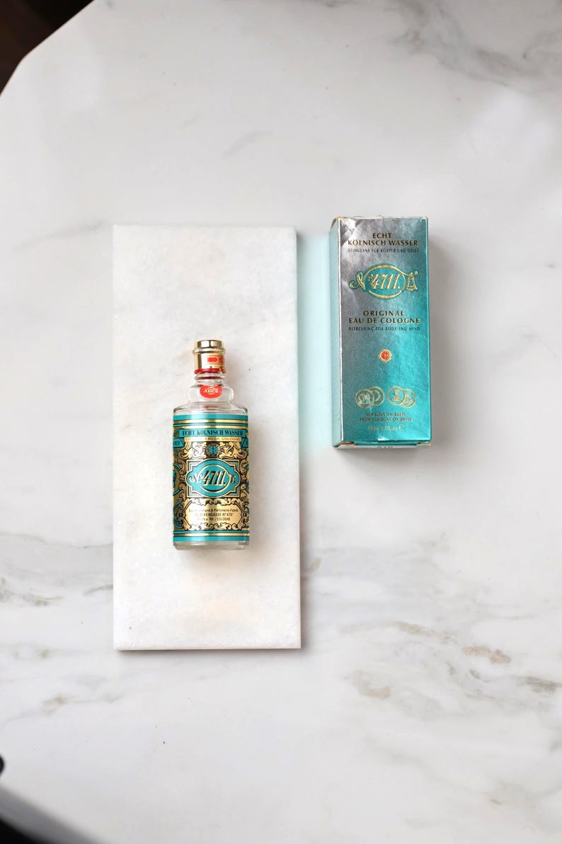 May include: A bottle of 4711 Original Eau de Cologne with a gold and teal label and a matching box. The bottle is on a white marble surface. The text on the bottle and box is in German and English.