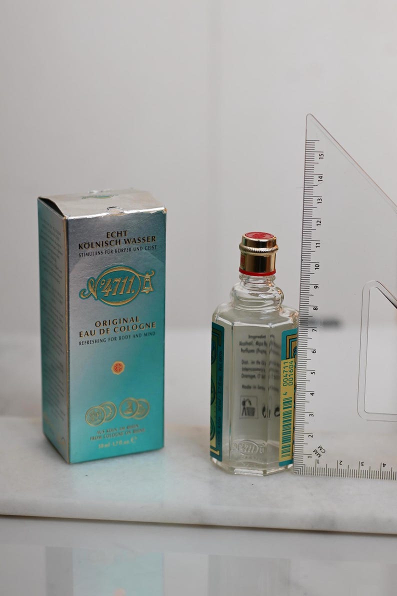 May include: A bottle of 4711 Original Eau de Cologne, with a red and gold cap, next to its teal and silver box. The bottle is approximately 3 inches tall. The box and bottle have text in German.