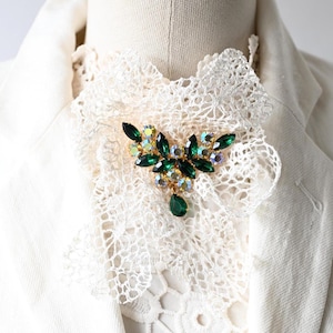 Op de afbeelding: Een vintage broche met smaragdgroene en iriserende kristallen, gezet in goudkleurig metaal. De broche heeft een bloemvormig ontwerp met een druppelvormige groene kristal. Het is vastgemaakt aan een witte kanten kraag.
