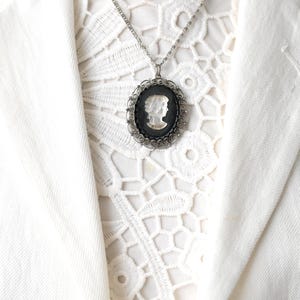 Op de afbeelding: Een zilverkleurige camee hangerketting. De ovale hanger heeft een wit profiel tegen een zwarte achtergrond, omlijst door een decoratieve zilveren rand. De ketting wordt getoond op een witte kanten top en een witte blazer.