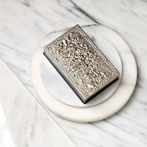 Peut inclure: Une boîte rectangulaire en métal argenté présentant un relief complexe sur le couvercle représentant plusieurs personnages en tenue historique, avec une base noire et une bordure décorative entourant la scène détaillée sur le dessus.