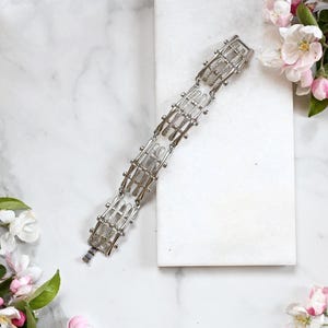 Op de afbeelding: Een zilverkleurige armband met een geometrisch ontwerp. De armband heeft rechthoekige schakels die zijn verbonden door afgeronde elementen. De schakels hebben een rasterpatroon. De armband wordt gepresenteerd op een wit oppervlak met roze en witte bloemen.