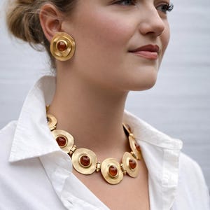 Può includere: Set di gioielli color oro con collana e orecchini abbinati. La collana presenta una serie di pendenti circolari con pietre color ambra. Gli orecchini sono grandi, circolari e presentano anche pietre color ambra. I gioielli sono indossati sopra una camicia bianca.