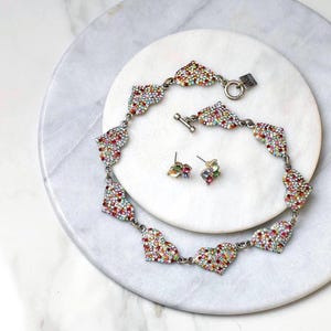 Op de afbeelding: Een zilverkleurige ketting en bijpassende oorbellen set. De ketting en oorbellen hebben hartvormige ontwerpen bezet met kleurrijke strass-steentjes. De sieraden worden gepresenteerd op een wit marmeren oppervlak.