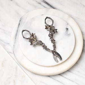 May include: A pair of silver-toned metal grape shears featuring intricate embossed detailing of grape clusters and vine leaves along the handles. The handles are shaped like twisted branches, and the blades are curved for cutting grapes.