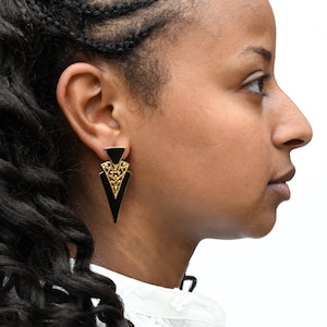 May include: A triangular earring featuring a black enamel top and bottom section separated by a central gold-toned metal piece with an intricate filigree cutout design. The earring has a sharp, pointed base and a polished, reflective finish.