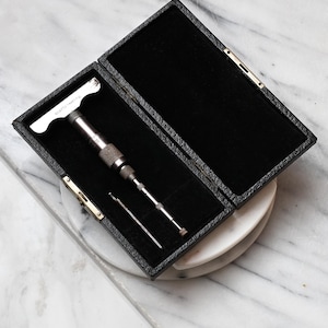 Puede incluir: Una herramienta de metal en forma de T con agarre moleteado y una varilla de metal delgada separada, alojada en un estuche rectangular negro texturizado con interior de terciopelo negro. La herramienta presenta marcas grabadas y un acabado metálico.