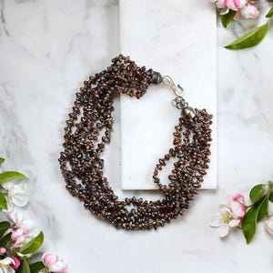 Op de afbeelding: Een ketting met meerdere strengen met kleine, donkerbruine kralen. De ketting is vastgemaakt met een zilverkleurige sluiting. De sieraden worden gepresenteerd op een wit marmeren oppervlak, met roze en witte bloemen op de achtergrond.