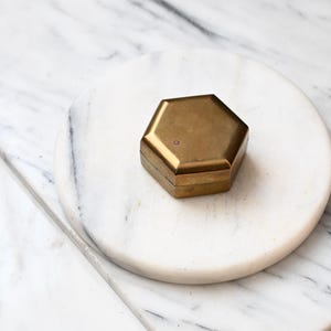 Puede incluir: Una pequeña caja hexagonal de latón con un acabado metálico liso y una tapa plana. El recipiente presenta una forma geométrica sencilla con bordes limpios y un color marrón dorado uniforme, mostrando una construcción sólida sin marcas visibles.