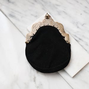Könnte beinhalten: Eine schwarze Geldbörse mit einem dekorativen silberfarbenen Verschluss. Der Verschluss hat ein verziertes Design mit geätzten Details und einem kleinen Knopf oben. Die Geldbörse ist aus einem weichen, dunklen Stoff gefertigt.