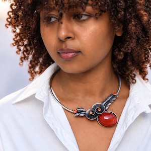 Op de afbeelding: Een zilverkleurige metalen choker-halsketting met een groot, glad, ovaalvormig oranje steen als middelpunt. Het ontwerp bevat metalen veerachtige accenten en kleine, ronde, feloranje ingelegde stenen langs de gebogen kraagstructuur.