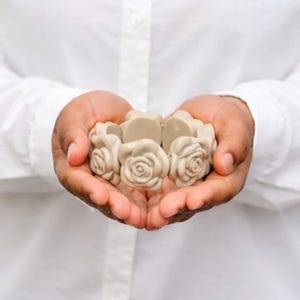 Op de afbeelding: Een armband met beige roosvormige kralen. De armband wordt in komvormige handen gehouden tegen een witte overhemdachtergrond. De rozen hebben gedetailleerde bloemblaadjes en een neutrale kleur.