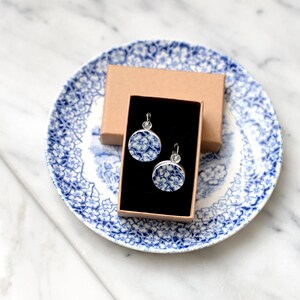 May include: A pair of round, blue and white floral earrings with silver-colored hooks, displayed in a black box inside a small brown box. The boxes rest on a blue and white patterned plate.