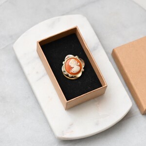 May include: A vintage cameo brooch with a carved woman's profile in white on an orange background, set in a gold-tone floral frame. The brooch is presented in a small, open, brown box with a black interior, on a white marble tray.