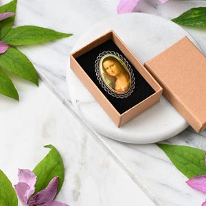 May include: An oval brooch featuring a reproduction of the Mona Lisa painting, set within a silver-toned metal frame with a twisted rope border. The brooch is presented inside a small, rectangular, light brown cardboard gift box with a black insert.