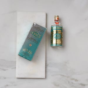 May include: A bottle of 4711 Original Eau de Cologne and its box, displayed on a white marble surface. The bottle is clear with a gold and teal label. The box is teal with gold lettering. The text "4711" is visible on both the bottle and the box.