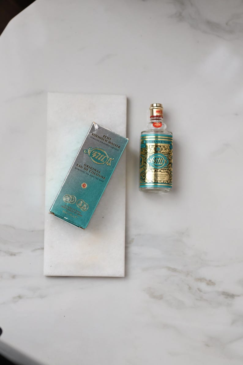 May include: A bottle of 4711 Original Eau de Cologne and its box, displayed on a white marble surface. The bottle is clear with a gold and teal label. The box is teal with gold lettering. The text "4711" is visible on both the bottle and the box.