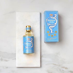 Pode incluir: Uma garrafa de "Boldoo! Eau de Cologne" com um rótulo azul claro e detalhes dourados, ao lado de sua caixa correspondente. A garrafa e a caixa apresentam um design de coroa e volutas decorativas. A garrafa está em uma bandeja de mármore branco.