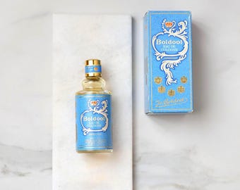 May include: A bottle of "Boldoo! Eau de Cologne" with a light blue label and gold accents, next to its matching box. The bottle and box feature a crown design and ornate scrollwork. The bottle is on a white marble tray.