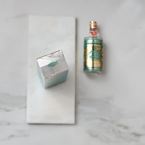 May include: An overhead shot of a perfume bottle and box on a white marble surface. The perfume bottle is gold and teal with a clear glass top. The box is silver with teal accents and a logo.