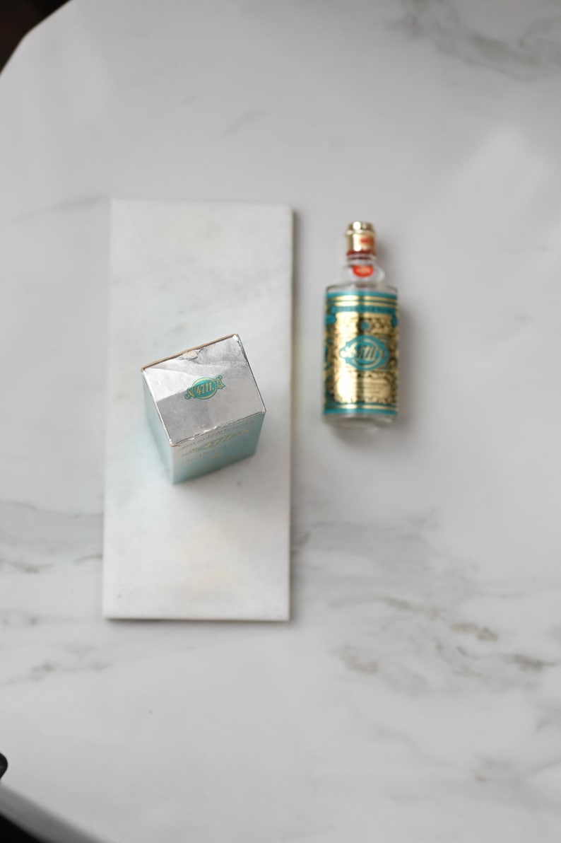 May include: An overhead shot of a perfume bottle and box on a white marble surface. The perfume bottle is gold and teal with a clear glass top. The box is silver with teal accents and a logo.