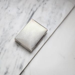 Op de afbeelding: Een rechthoekige zilverkleurige metalen doos met een fijne, verhoogde ruitstructuur over het gehele bovenoppervlak. Het object heeft strakke, rechte randen en een metallic afwerking die het licht op het rasterpatroon weerkaatst.