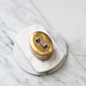 Pode incluir: Uma caixa oval de latão com tampa articulada apresentando um fecho central de metal em forma de laço. A caixa tem um acabamento dourado polido com sinais visíveis de envelhecimento e pátina, sobre uma superfície de mármore branco liso.