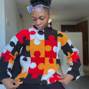 Puede incluir: Un colorido suéter de ganchillo con un diseño de rompecabezas. El suéter combina piezas de rompecabezas rojas, negras, blancas y amarillas. Tiene cuello alto y mangas largas. Este suéter hecho a mano es una prenda única y llamativa.