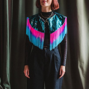 May include: A teal sequined vest with vibrant pink and blue fringe. The vest is worn over a black long-sleeved top and black trousers. The fringe is a gradient of pink to blue. A gold necklace is also visible.