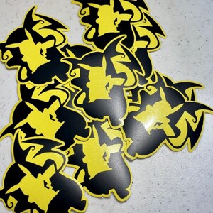 May include: A collection of yellow and black die-cut stickers featuring a Pikachu silhouette within a lightning bolt design. The stickers have a glossy finish and are scattered across a light-colored surface. The stickers are likely for fans of the popular animated series.