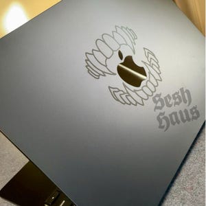 May include: A dark gray laptop with a silver graphic of an apple with wings and the words "Sesh Haus" in a gothic font. The laptop is open, revealing the keyboard.