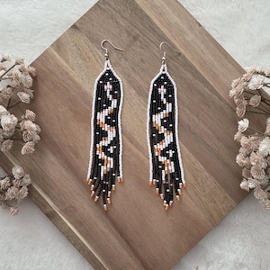 May include: A pair of long, dangling earrings crafted from small seed beads in black, white, and orange. The beads are arranged in a geometric, zigzag pattern that runs vertically down the length of each earring, finished with silver-toned hooks.