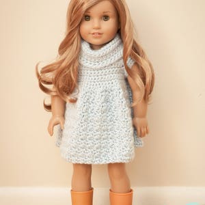 May include: A doll with long, wavy blonde hair wearing a light blue, crocheted dress and orange boots. The doll is standing on a wooden floor against a cream-colored wall. The dress has a cowl neck.