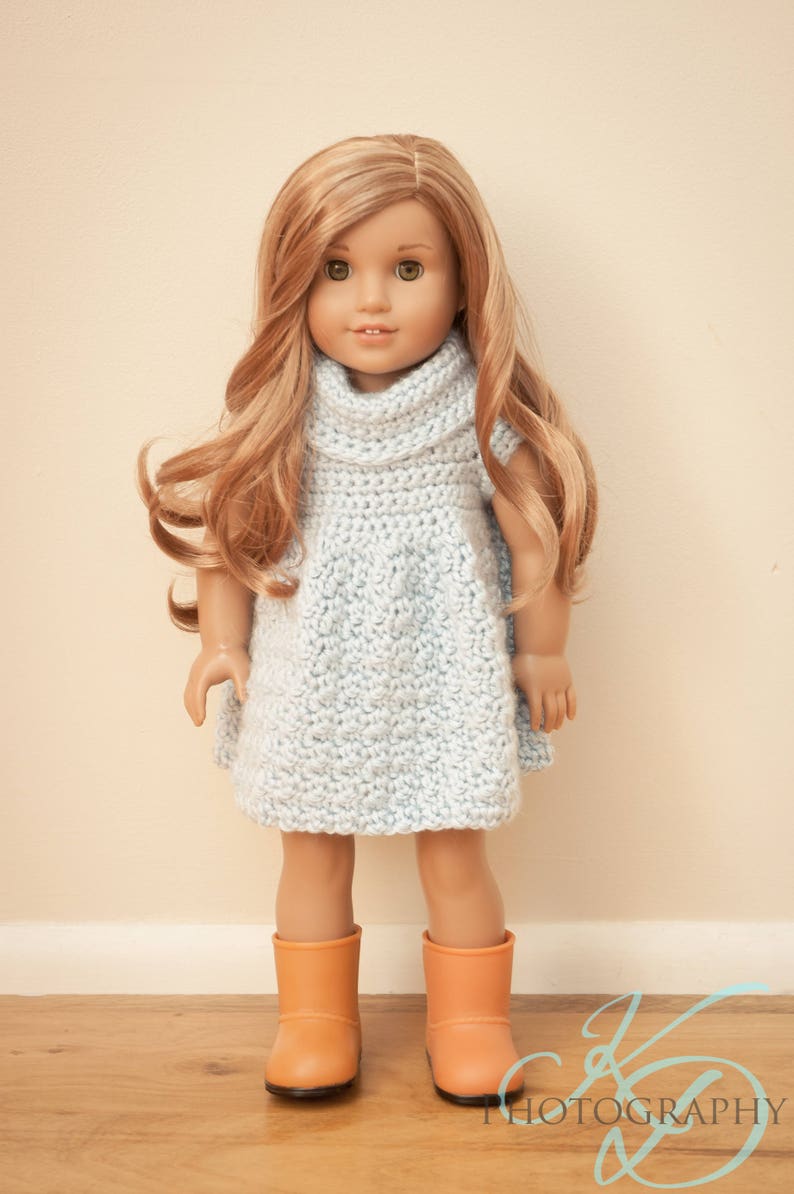 May include: A doll with long, wavy blonde hair wearing a light blue, crocheted dress and orange boots. The doll is standing on a wooden floor against a cream-colored wall. The dress has a cowl neck.