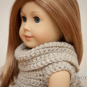 May include: A doll with long, light brown hair and blue eyes, wearing a gray, hand-knitted sweater. The sweater has a high, rolled collar. The doll's skin is a pale tone, and it has a slight smile. The background is a soft, neutral color.