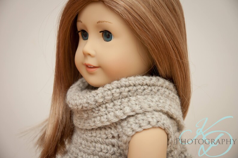 May include: A doll with long, light brown hair and blue eyes, wearing a gray, hand-knitted sweater. The sweater has a high, rolled collar. The doll's skin is a pale tone, and it has a slight smile. The background is a soft, neutral color.