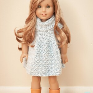 May include: A doll wearing a light blue, crocheted dress with a cowl neck and short sleeves. The doll has long, wavy blonde hair and is wearing orange boots. The doll is standing on a wooden surface.