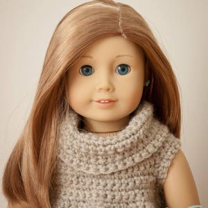 May include: A doll with long, light brown hair and blue eyes, wearing a light gray, crocheted turtleneck sweater. The doll has a gentle smile and small, blue earrings. The background is a solid, neutral color.