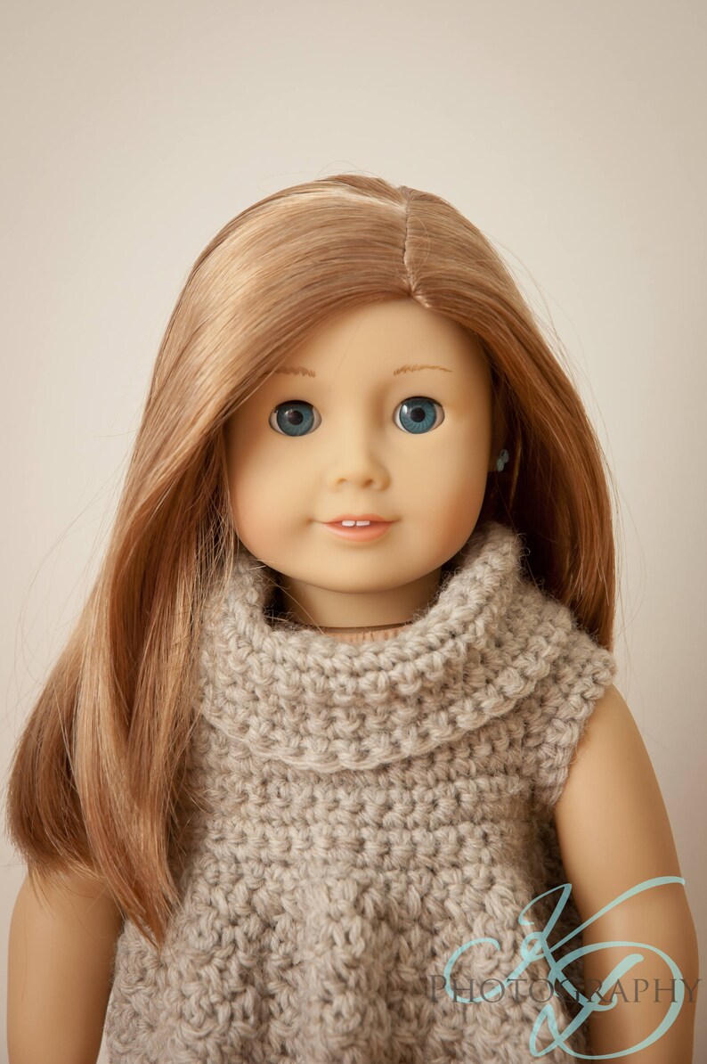 May include: A doll with long, light brown hair and blue eyes, wearing a light gray, crocheted turtleneck sweater. The doll has a gentle smile and small, blue earrings. The background is a solid, neutral color.