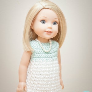 May include: A doll wearing a crocheted dress with a light blue top and white bottom. The doll has blonde hair and blue eyes and is wearing a pearl necklace.