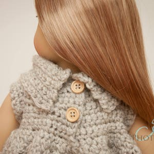 May include: A light gray, crocheted doll vest with two wooden buttons. The vest has a scalloped collar and is worn by a doll with long, light brown hair. The vest is handmade and suitable for a doll.