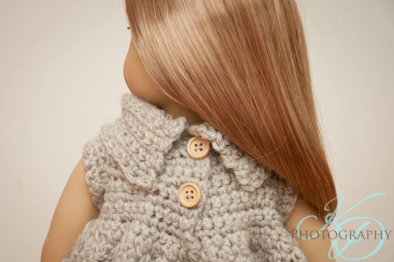 May include: A light gray, crocheted doll vest with two wooden buttons. The vest has a scalloped collar and is worn by a doll with long, light brown hair. The vest is handmade and suitable for a doll.