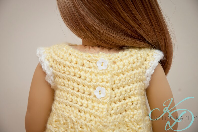 Op de afbeelding: Een gele, gebreide, mouwloze jurk met witte bies en twee witte, bloemvormige knopen op de rug.