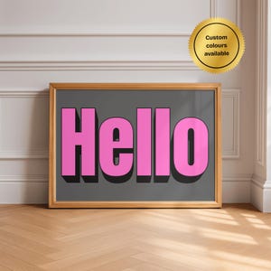 Puede incluir: Una pieza de arte mural rectangular enmarcada que presenta la palabra Hello en letras rosas llamativas de estilo 3D sobre un fondo gris oscuro. Un sello circular dorado en la esquina superior derecha contiene el texto Custom colours available.