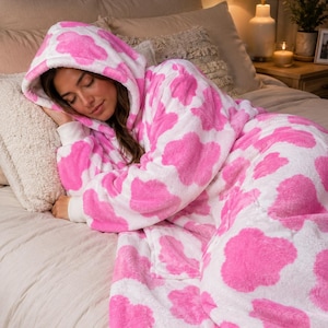 May include: A plush, hooded robe featuring a white base with a repeating pattern of bright pink, cloud-like shapes. The garment is made of a soft, fuzzy material and includes long sleeves with white cuffs and a hood that covers the head.